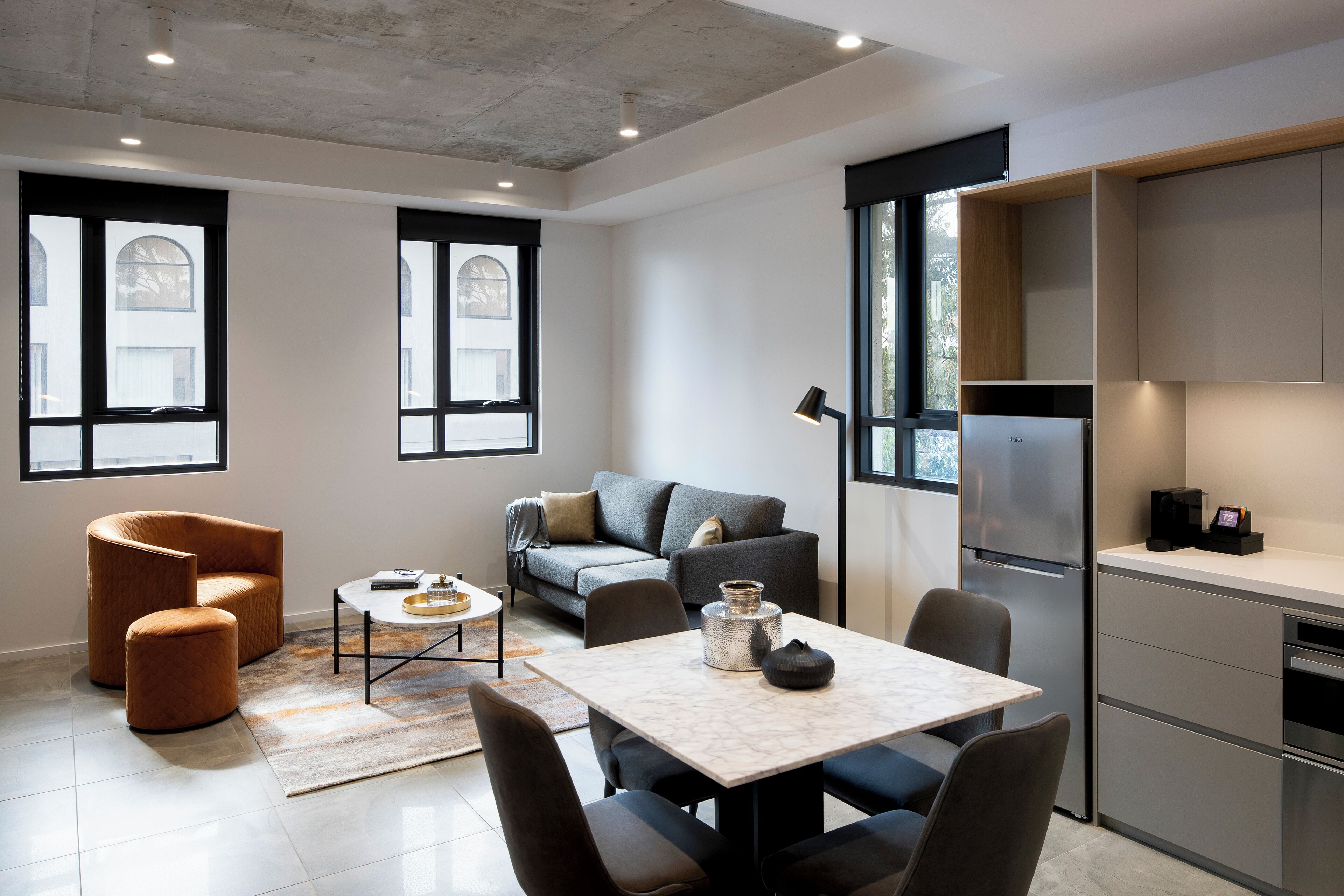 Una sala moderna con sofá, sillón y mesa de centro. Se observa un área de cocina con refrigerador y armarios.