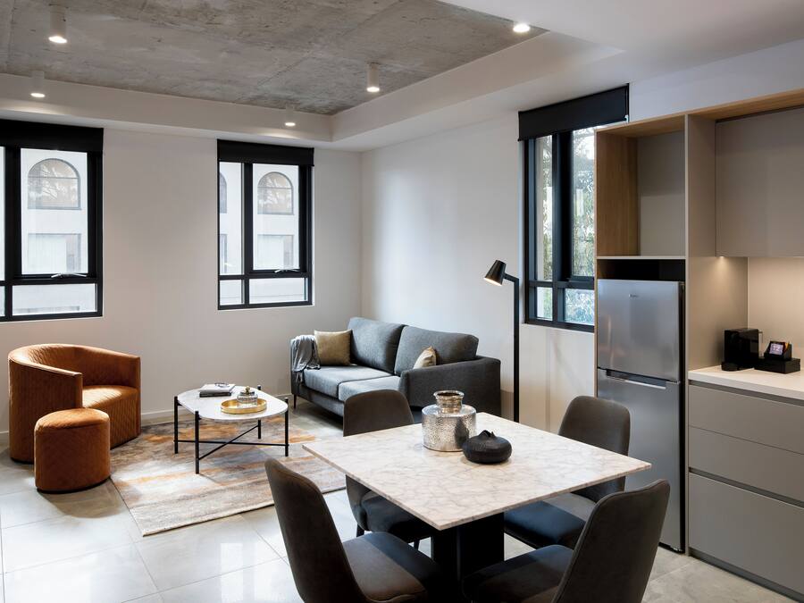 A modern living room with a sofa, armchair, and coffee table. There is a kitchen area with a refrigerator and cabinets.