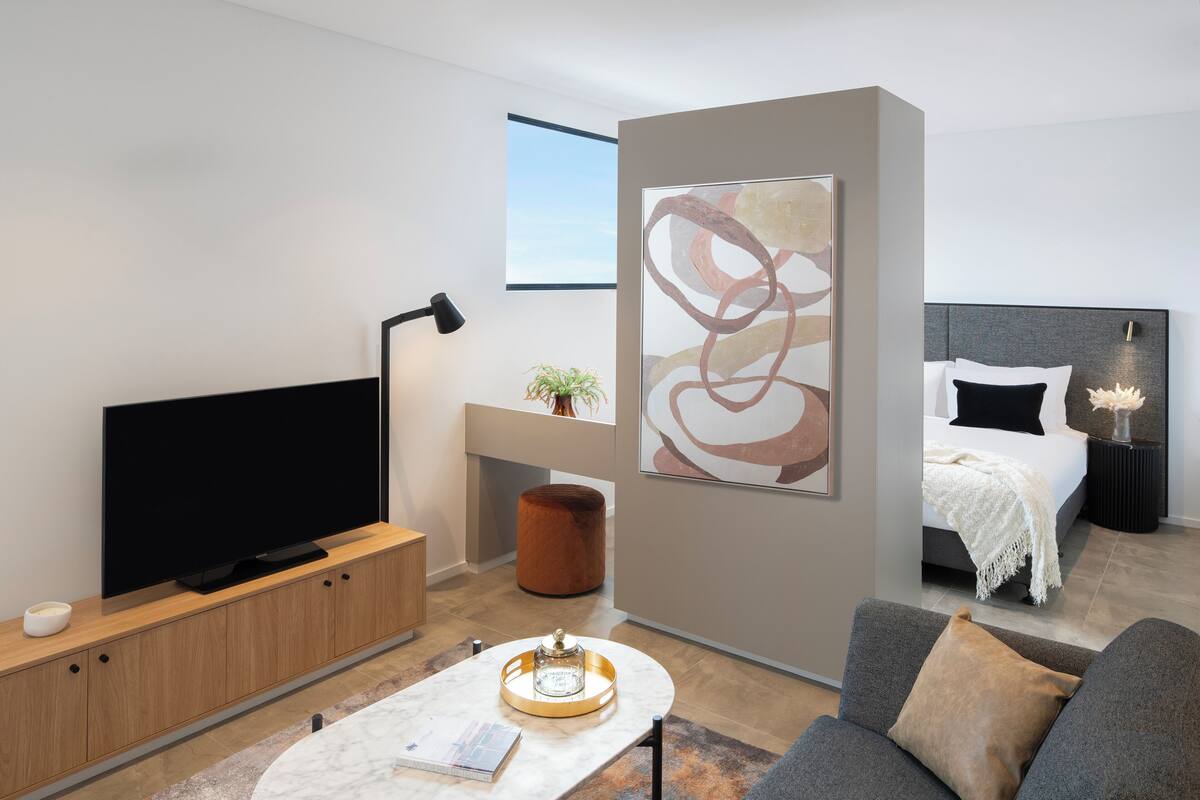 Ein modernes Hotelzimmer mit Flachbildfernseher, einer Couch, einem Couchtisch, einem Bett mit Kopfteil, einem Nachttisch und einem großen abstrakten Gemälde an der Wand.