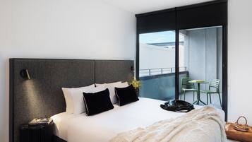 A modern hotel room with a large bed, a bedside table, a lamp, a view of the balcony, and a chair outside.
