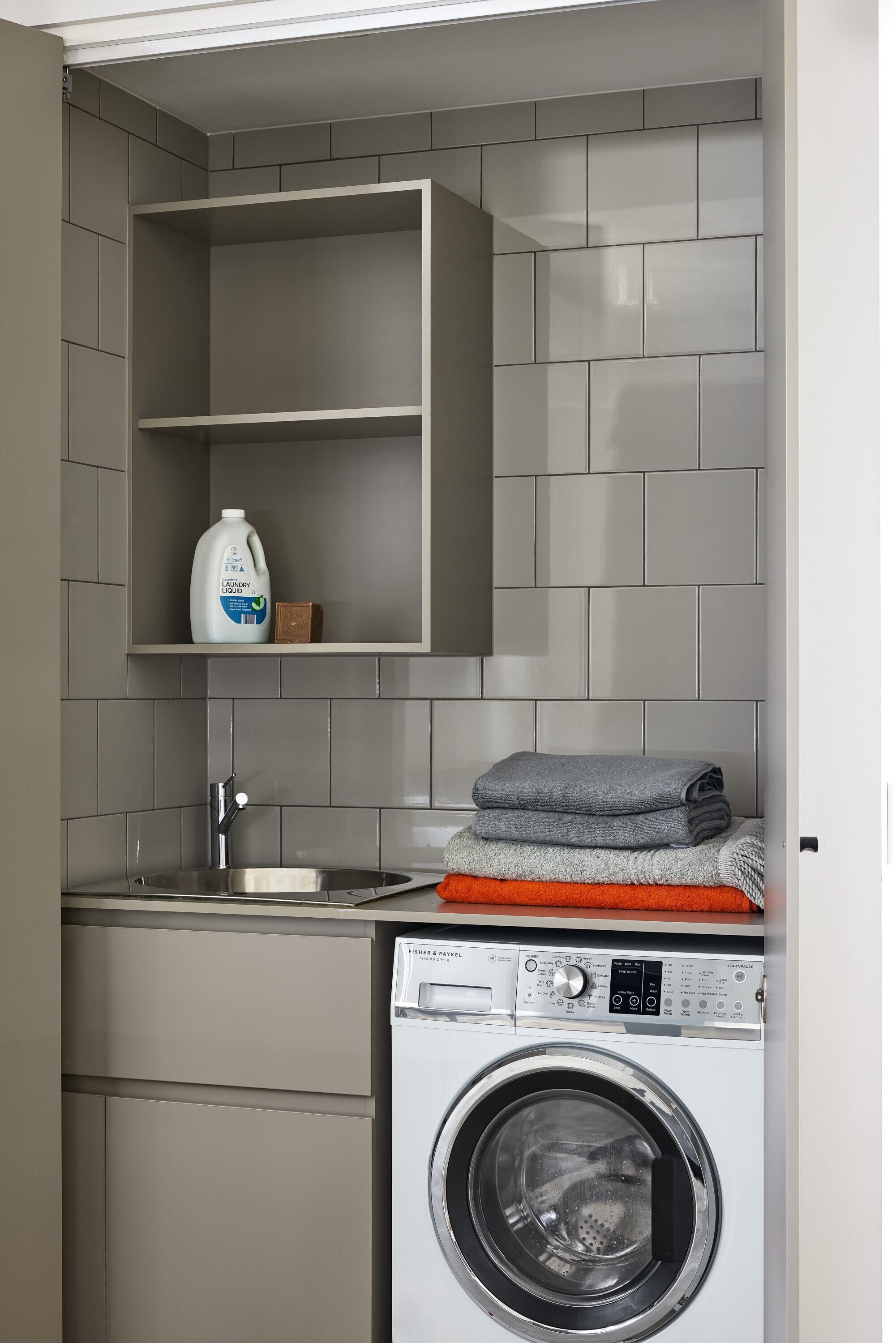 A compact laundry area with a washing machine, a shelf with a detergent bottle, and a folded towel on top.