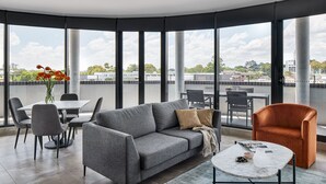 A modern living room with a grey sofa, a coffee table, and a dining area with a table and chairs. Large windows offer a view of the cityscape. - Punthill Parramatta (Parramatta)