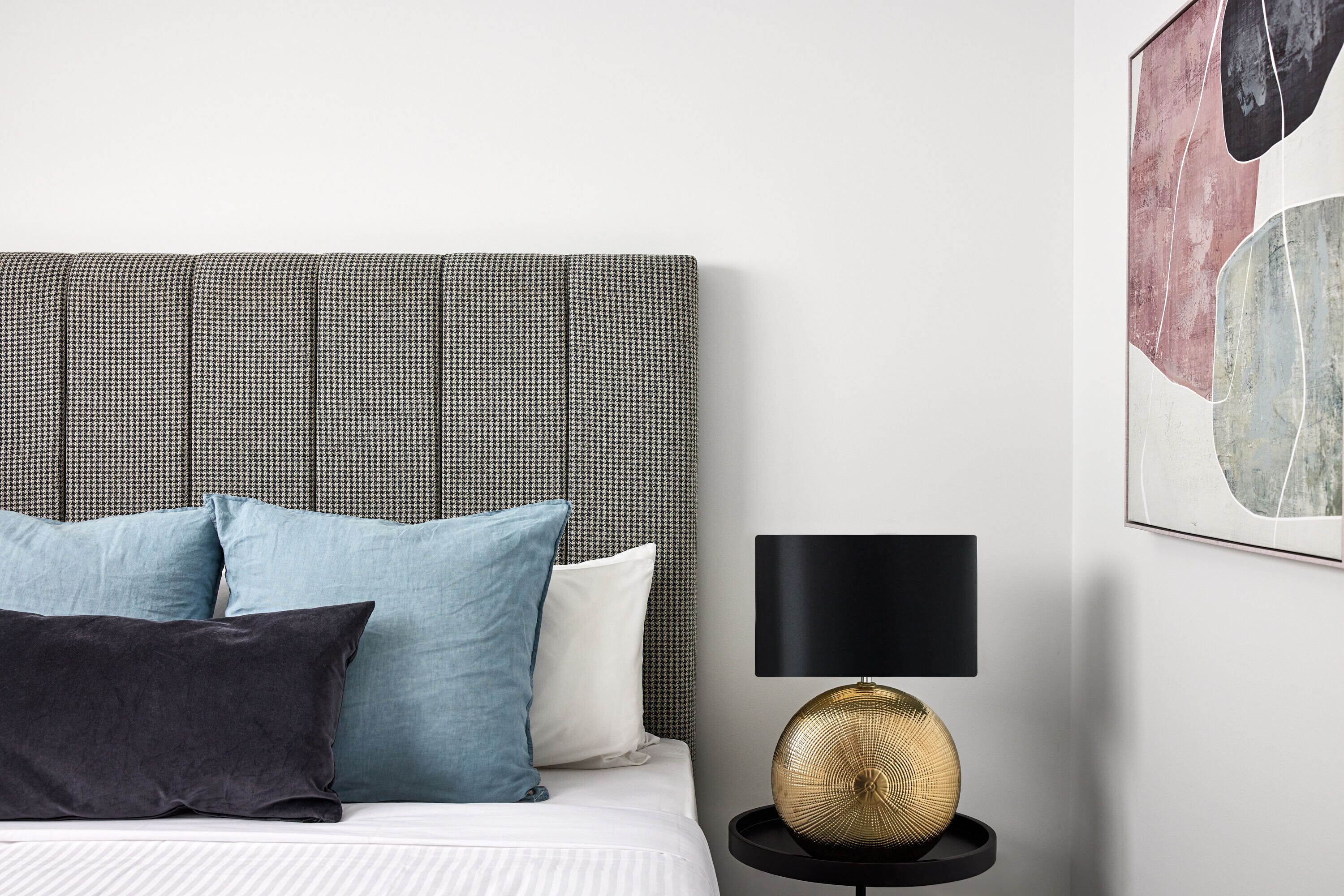 A neatly made bed with a checkered headboard, a black lamp, and a framed abstract painting on the wall.