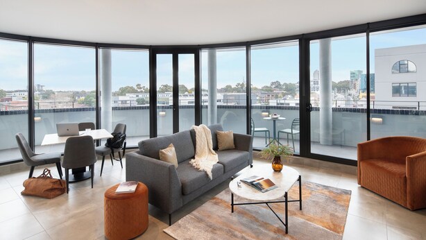 A modern living room with a grey sofa, two armchairs, a coffee table, and a view of the city through large windows. - Punthill Parramatta (Parramatta)