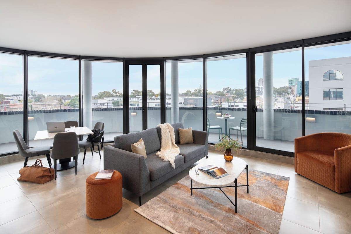 Ein modernes Wohnzimmer mit einem grauen Sofa, zwei Sesseln, einem Couchtisch und Blick auf die Stadt durch große Fenster.