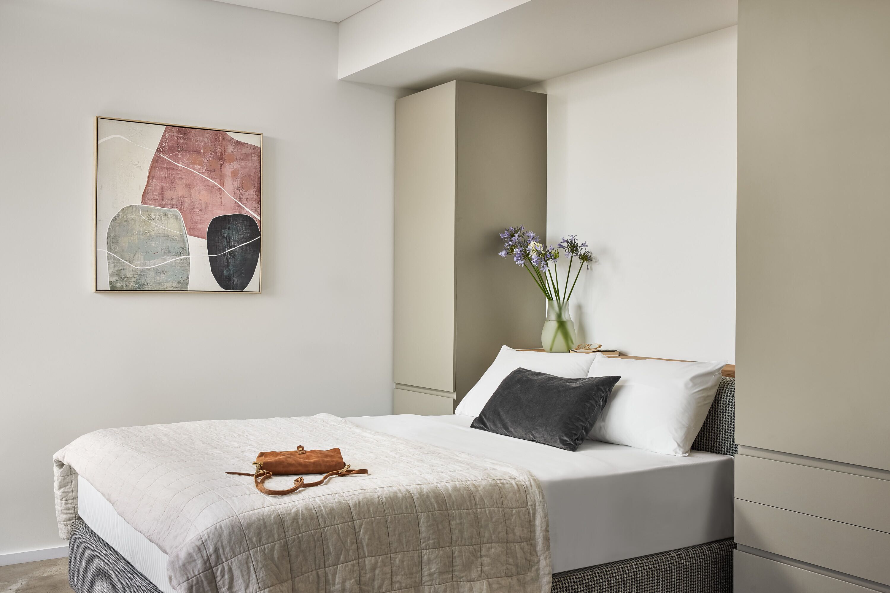 A neatly arranged bedroom with a bed, a bedside table, a vase with flowers, and a painting on the wall.