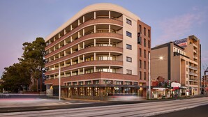 A multi-story hotel with a curved facade, multiple balconies, and ground-level commercial spaces. - Punthill Parramatta (Parramatta)