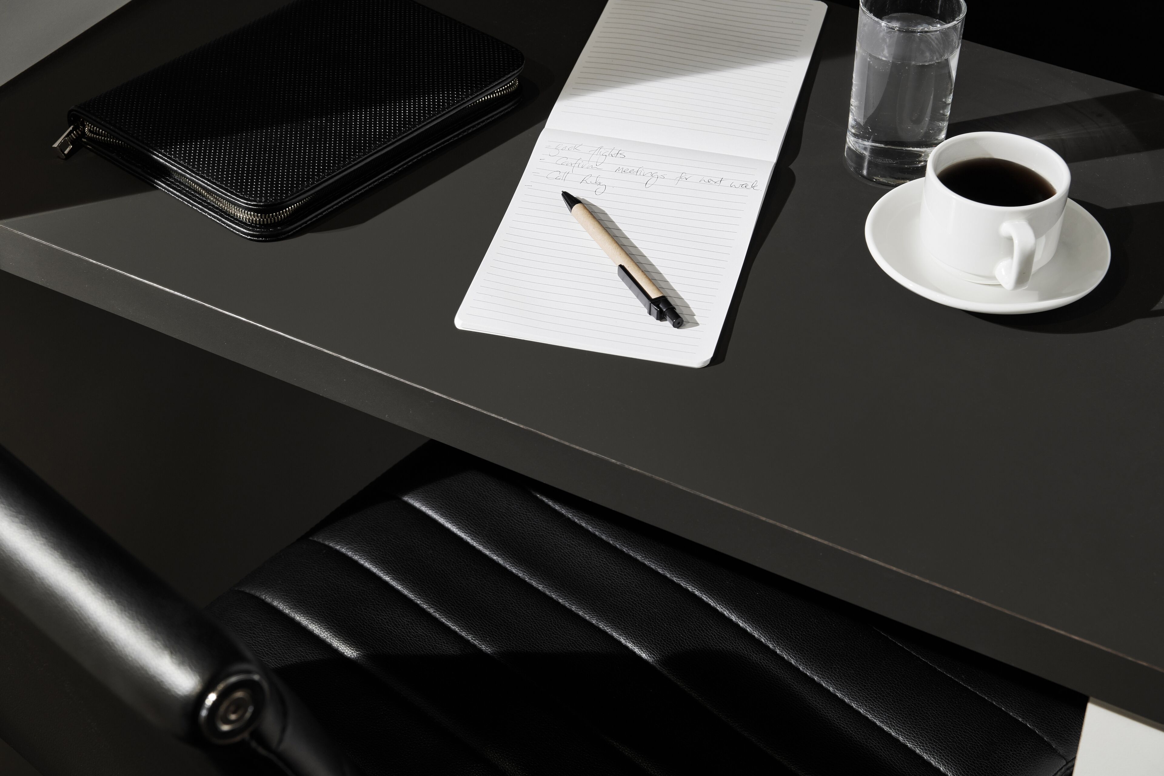 Ein schwarzer Ledersessel, ein Notizbuch mit Stift, eine Kaffeetasse und eine Wasserflasche auf einem Tisch.