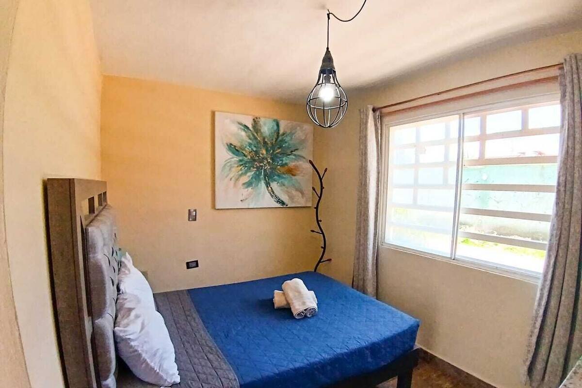 A small, neatly arranged bedroom with a bed, a painting, a window with curtains, and a hanging light fixture.