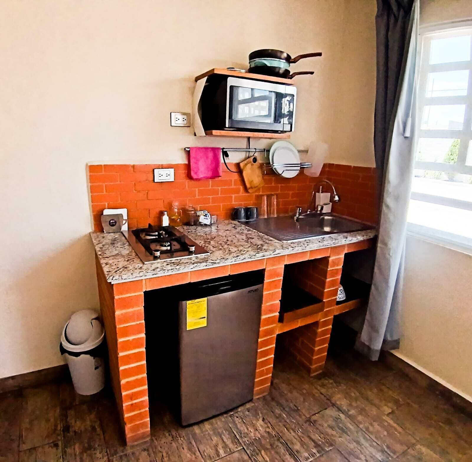 Cocina compacta con estufa, lavamanos y microondas integrados. Cuenta con un revoque de ladrillo y un refrigerador pequeño.
