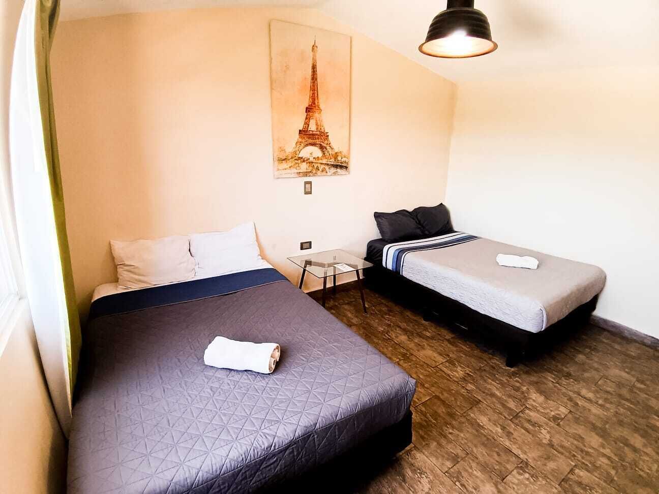 Une chambre d’hôtel avec deux lits, une petite table, une lampe et un tableau représentant la Tour Eiffel.