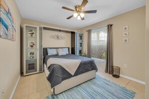 Une chambre à coucher avec un lit, un ventilateur de plafond, une fenêtre avec des rideaux et un mur décoré de tableaux encadrés.