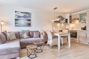 A modern living room with a sofa, a dining table, and chairs. There is a kitchen area with appliances and a hanging light fixture. - Hansa-residenz II Appartement 9 - Hansa Residenz II (Scharbeutz)