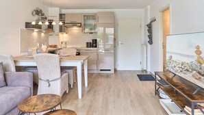 Une cuisine moderne avec des meubles blancs, un coin repas avec une table et des chaises, et un salon avec un canapé et un téléviseur.