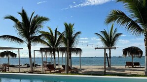 Un área de piscina frente a la playa, con tumbonas y palmeras.