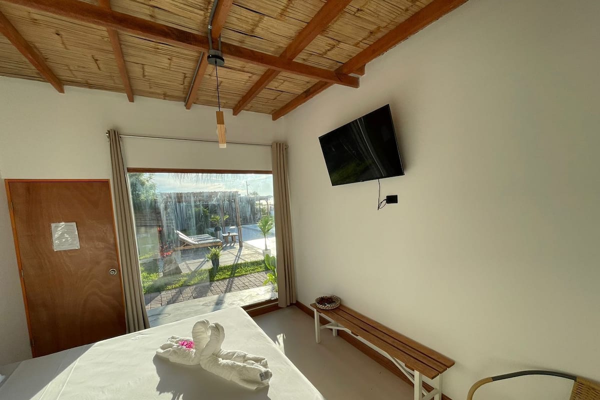 Ein Hotelzimmer mit Bett, einer Bank, einem Korbsessel, einem Fernseher an der Wand und Blick ins Freie.