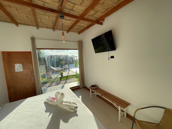 Habitación de hotel con cama, un banco, una silla de mimbre, televisor en la pared y vistas al exterior.
