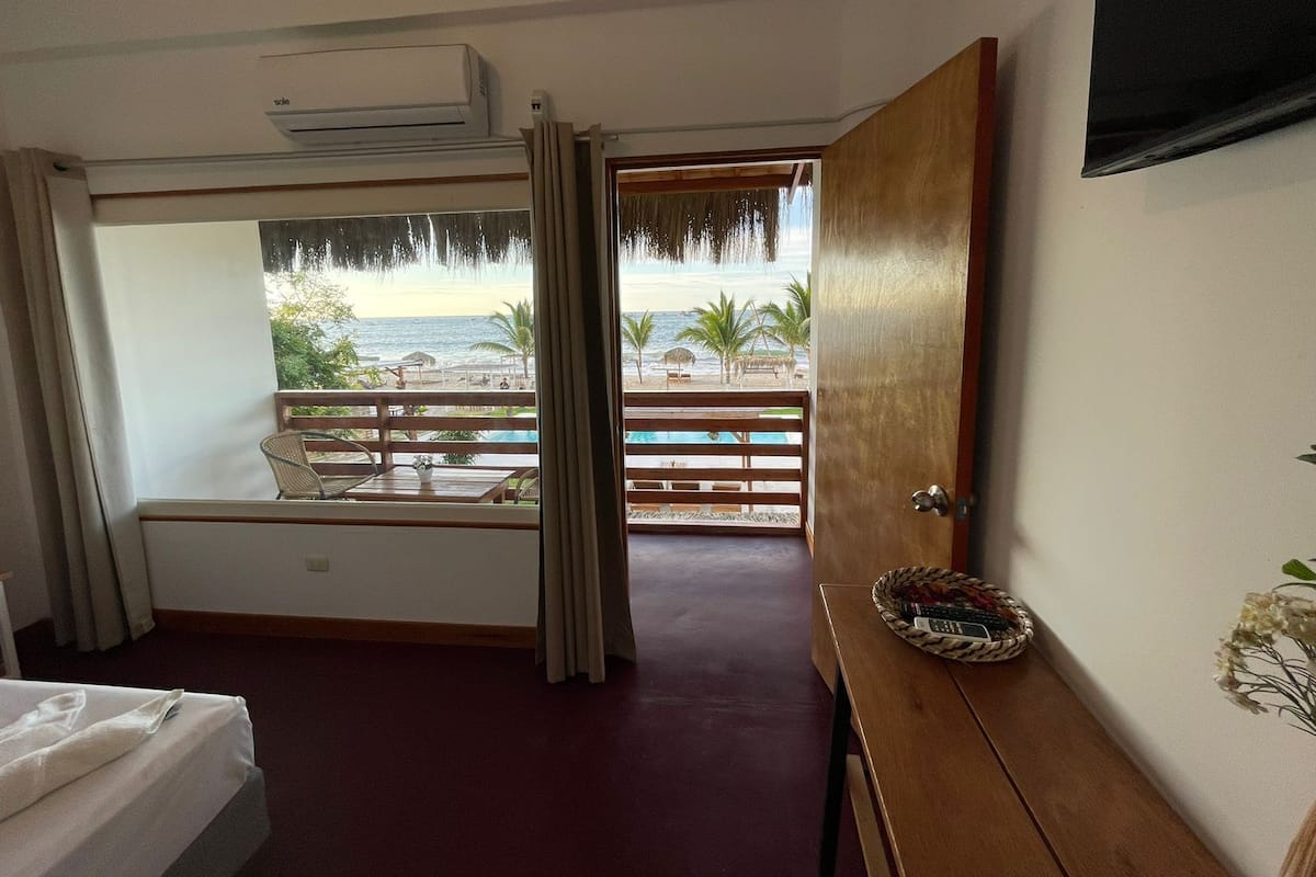 Ein Hotelzimmer mit Balkon und Blick auf einen Strand, einem Bett, einem Holztisch und einem Fenster mit Vorhängen.