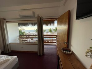 A hotel room with a balcony overlooking a beach, a bed, a wooden desk, and a window with curtains.