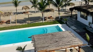 Zona de piscina con vista a la playa y al océano, rodeada de palmeras y estructuras de techo de paja.