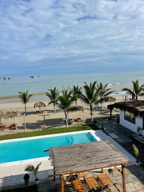 A pool area with a view of the beach and ocean, surrounded by palm trees and thatched-roof structures.