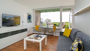 A modern living room with a television, a sofa with a yellow pillow, and a dining area with chairs and a table.