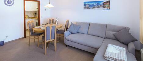 Ein Wohnzimmer mit grauem Sofa, einem Esstisch mit Stühlen, einer Hängelampe und einem Gemälde einer verschneiten Berglandschaft.