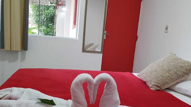Habitación de hotel con colcha roja, una toalla dispuesta en forma de corazón, almohada color crema y ventana con vistas a follaje.