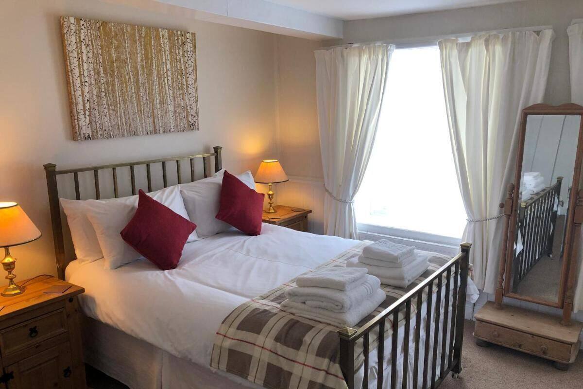 Una cama bien tendida con sábanas blancas y almohadas rojas, una lámpara de noche, una toalla doblada sobre la cama, una mesita de noche de madera, un espejo y una ventana con cortinas.