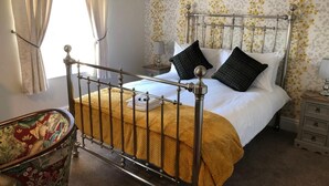 A bedroom with a metal bed frame, a yellow bedspread, two black pillows, a bedside table with a lamp, and a floral wallpaper. - the black swan (Leyburn)