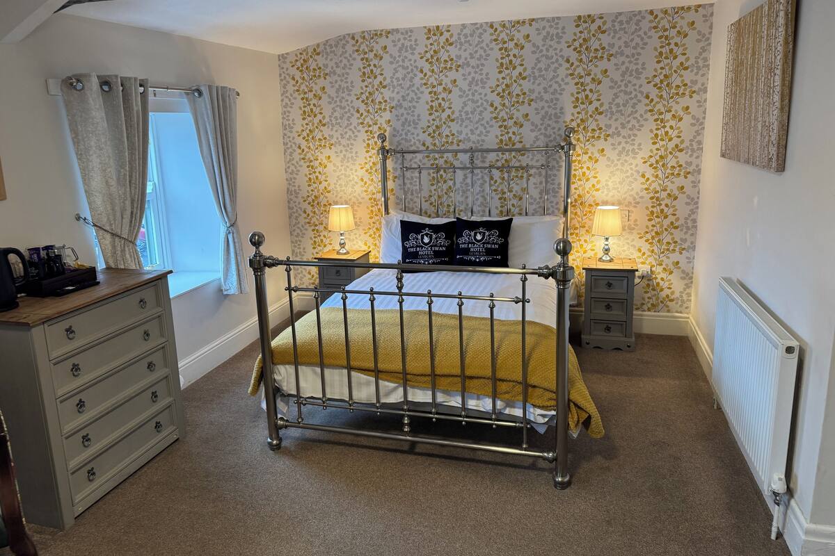 Un dormitorio con estructura de cama de metal, cómoda de madera, mesita de noche con lámpara y radiador.