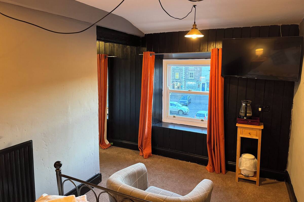 Una habitación con ventana, una silla, una mesita con hervidor y un televisor en la pared.