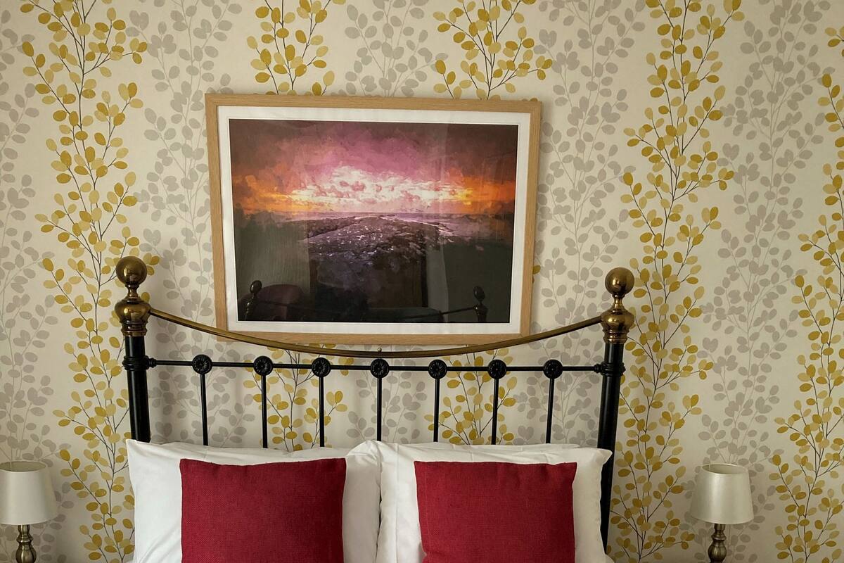 Una cama bien hecha con almohadas rojas y blancas, un cuadro enmarcado encima y una mesita de noche con una lámpara y un libro.
