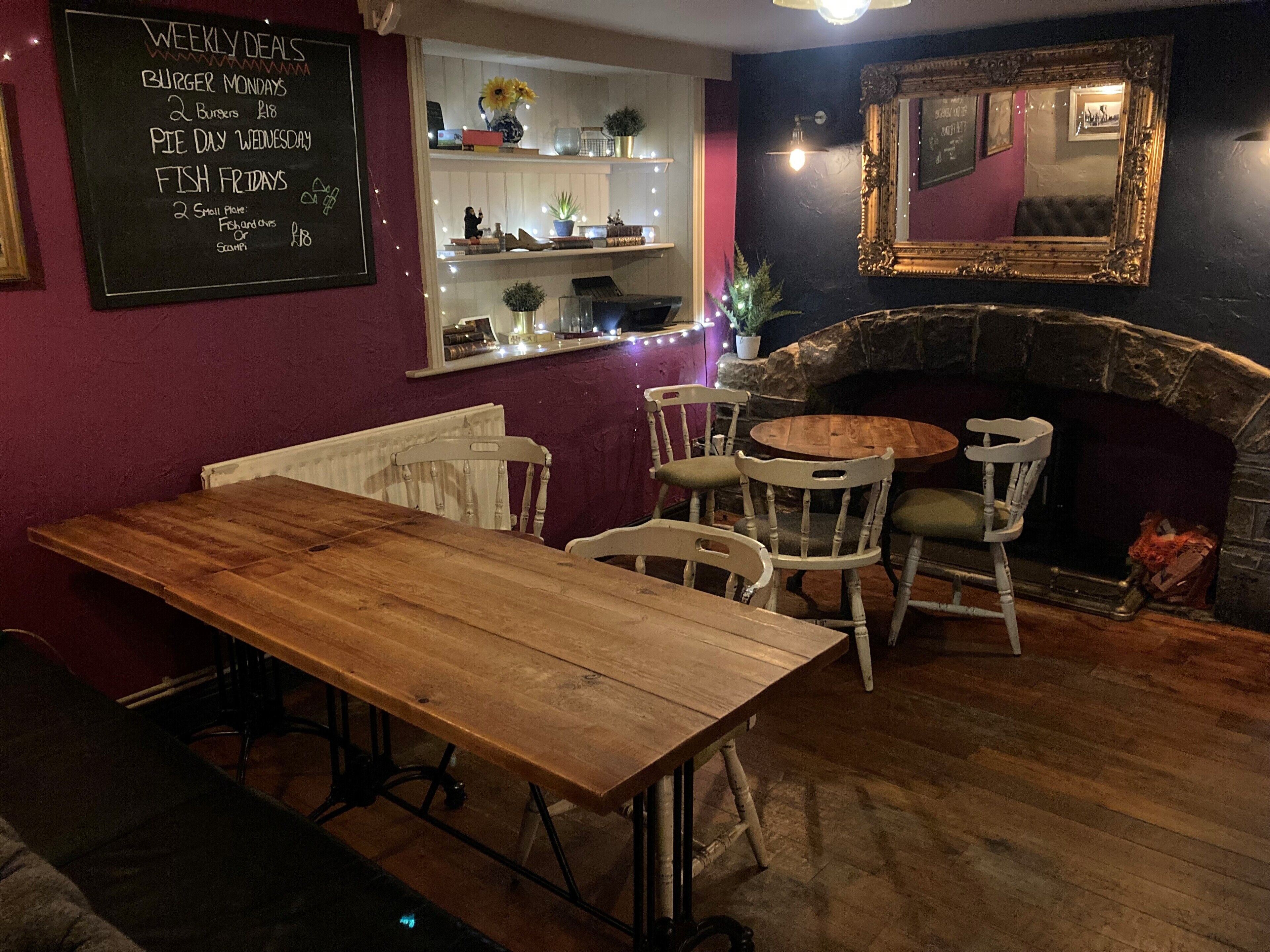 A cozy pub with a wooden table and chairs, a fireplace, and a chalkboard menu listing weekly deals.