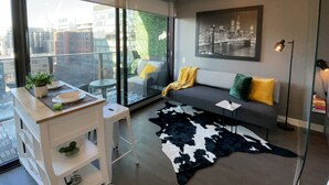Un salon moderne avec une cloison en verre, un canapé avec des coussins, un tapis noir et blanc, une petite table avec une plante et un tableau représentant un paysage urbain accroché au mur.