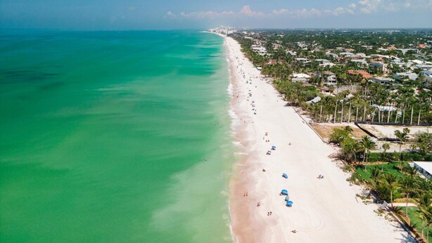 An aerial view of a beach with a clear turquoise sea, a sandy shore, and a residential area with houses and palm trees. - NAPLES GEM (Naples)