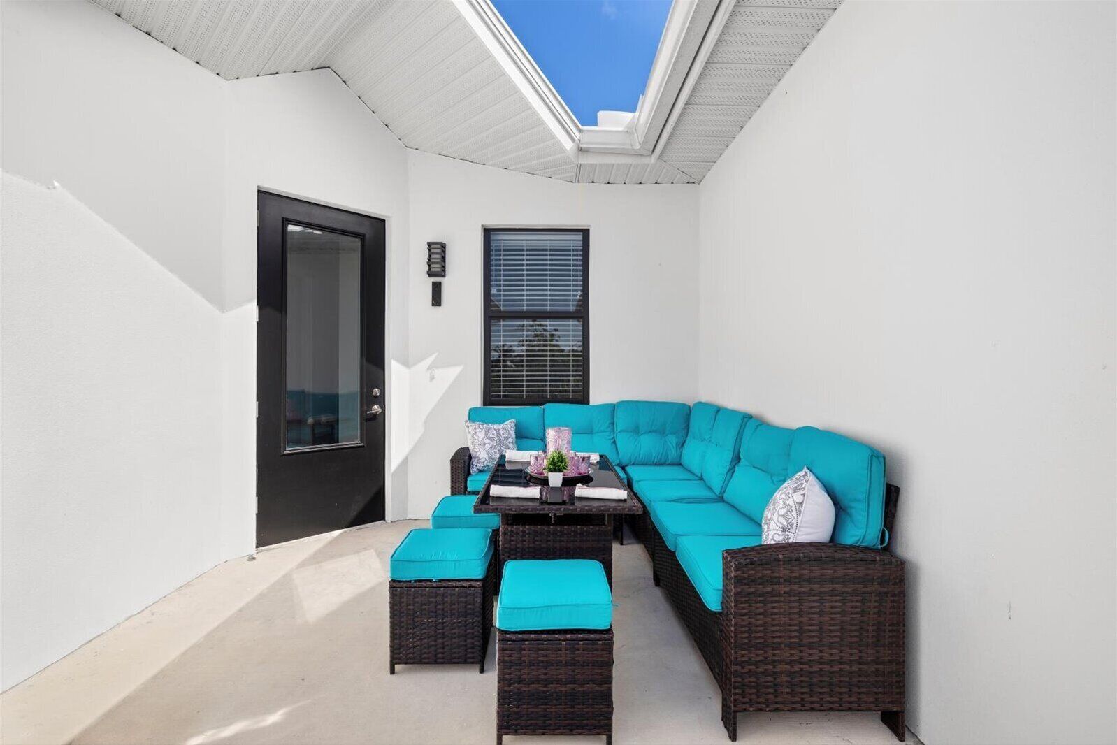 A sunlit patio with a teal sectional sofa, two ottomans, a small table, and a black door.
