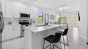 A modern kitchen with white cabinetry, a central island with bar stools, a microwave, oven, and a toaster. - NAPLES GEM (Naples)