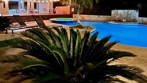 Vue nocturne d’un espace piscine avec des chaises longues et un palmier.