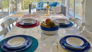 Un coin repas avec une table dressée pour deux personnes, une bouteille de vin et des fruits. La pièce est dotée de grandes fenêtres offrant une vue sur l’océan.