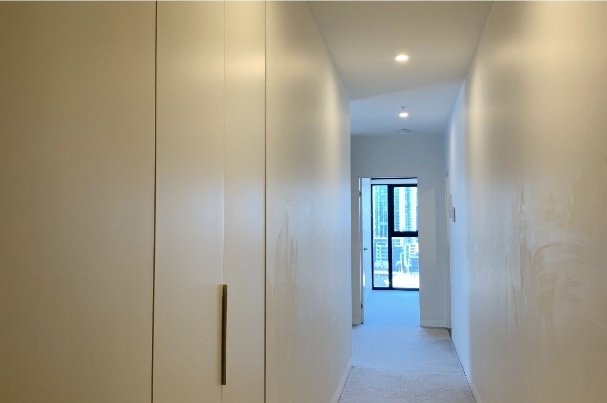 Un pasillo estrecho con paredes blancas y una puerta al final que da a una habitación luminosa con un ventanal grande.