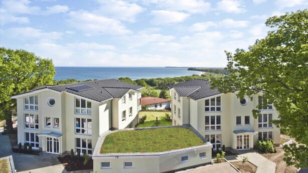 A multi-story residential building with a flat roof and a green roof section, surrounded by trees and a view of the sea. - Meeresblick Residenzen Fewo 67 Deluxe: Balkon, Meerblick, Sauna- u. Schwimmbadnutzung (Göhren)
