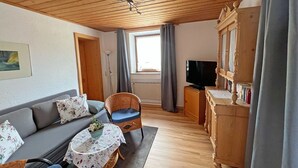 A cozy living room with a wooden ceiling, a sofa, a wicker chair, a small table with a floral tablecloth, and a television on a wooden cabinet.