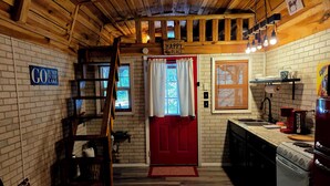 Interior acogedor de una cabaña con puerta roja, paredes de madera, chimenea de ladrillo, un altillo con escalera y una zona de cocina con estufa y lavaplatos.