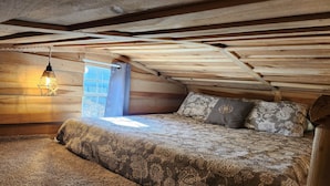 Un dormitorio acogedor en el ático con techo inclinado de madera, una cama con ropa de cama estampada, una lámpara colgante y una ventana con cortinas.
