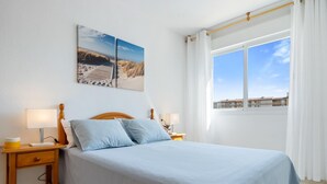 Un dormitorio con una cama, dos mesitas de noche, una mesilla auxiliar, una ventana con cortinas y vistas a edificios.