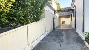 A narrow concrete pathway leading to a building entrance, flanked by a white fence and greenery. - Sydney’s 4BDR with large private yard / Eat-Sleep-Shop-Transit (Campsie)