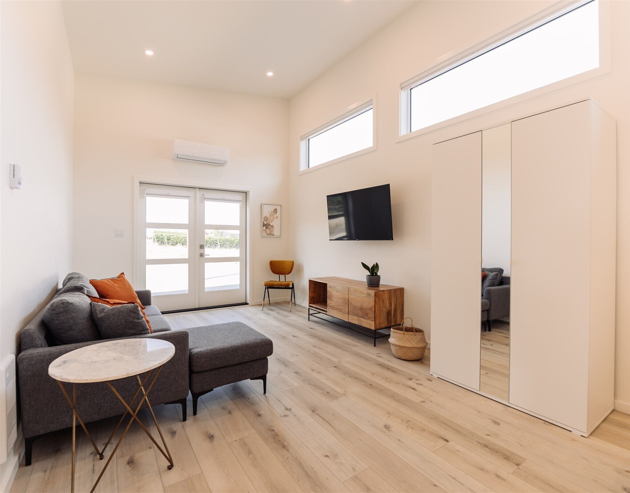 Un salon spacieux avec un canapé gris, une petite table ronde, un meuble en bois et un téléviseur fixé au mur.