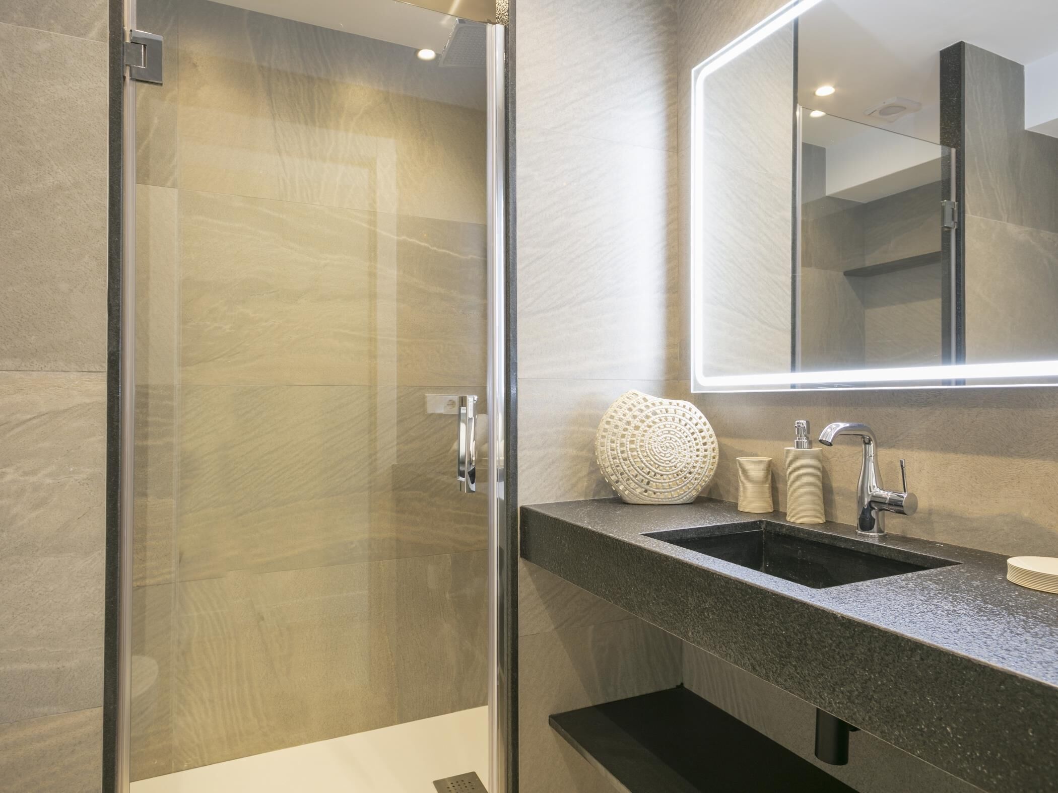 A modern bathroom with a glass shower enclosure, a granite countertop, a sink with a mirror, and a decorative vase.