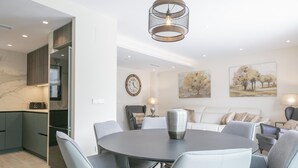 Ein modernes Wohnzimmer mit einem runden Esstisch, grauen Stühlen, einem grauen Sofa und einem Couchtisch. Eine Küchenzeile mit dunklen Schränken und einer Marmor-Spritzschutzwand ist ebenfalls zu sehen. Eine große Wanduhr und zwei Gemälde sind sichtbar.